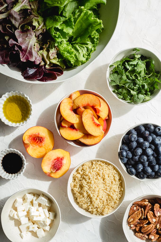Ingredients to make blueberry peach salad arranged in individuals bowls on a white surface.