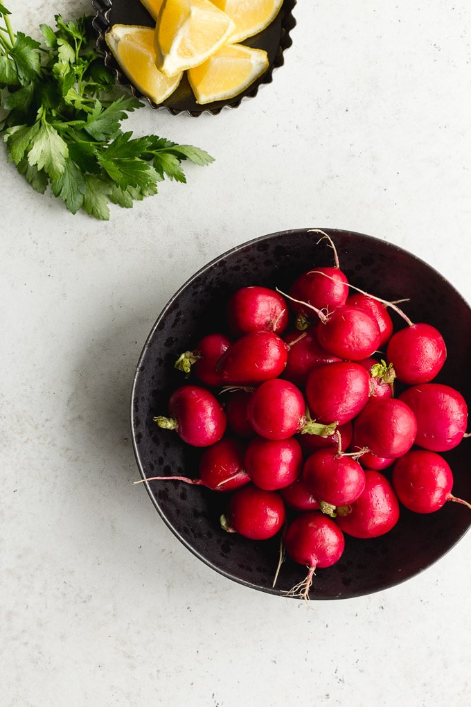 Whole radishes in a black bowl next to fresh parsley and lemon wedges.