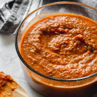 Homemade marinara sauce in a glass bowl next to a wooden spoon with sauce on it.