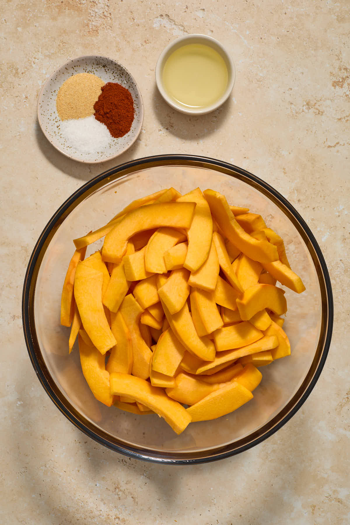 Pumpkin slices in a large glass bowl with oil and seasonings on the side.