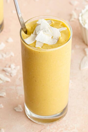 Angled view of a mango avocado smoothie in a tall glass with coconut slices on top.