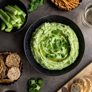 Edamame hummus served in a black bowl with crackers, bread and pretzels arranged around it.