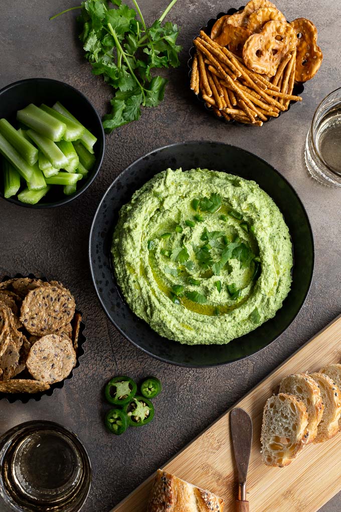 Overhead view of edamame hummus in a black bowl, surrounded by bowls of celery, crackers and pretzels.