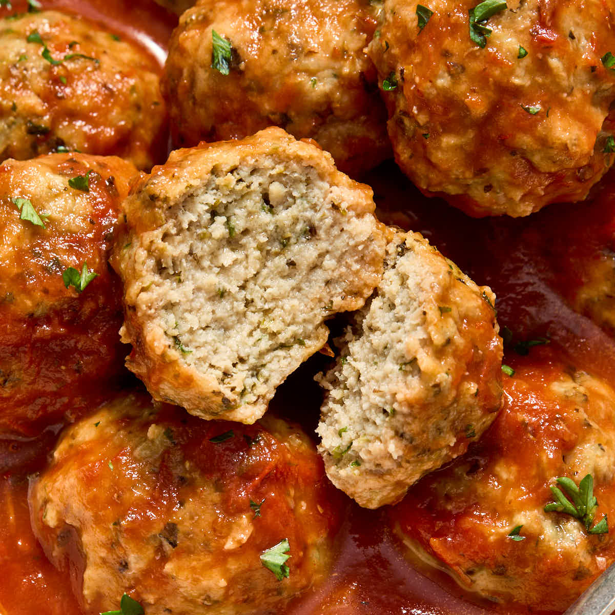 Close up of a chicken meatball cut in half on top of more meatballs topped with marinara.