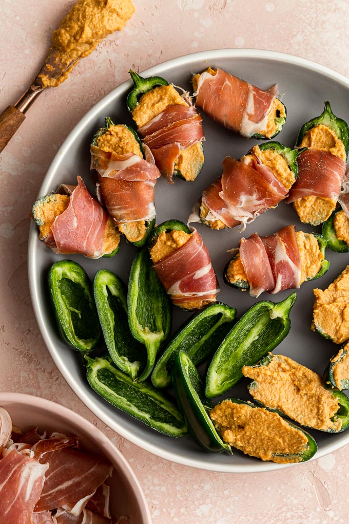 Jalapeño halves arranged on a plate, being stuffed with cashew cheese and wrapped in prosciutto.