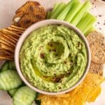 Overhead of arugula hummus in a pink bowl with chips, crackers, pretzels and veggies surrounding it.