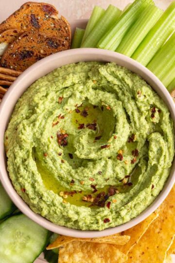 Overhead of arugula hummus in a pink bowl with chips, crackers, pretzels and veggies surrounding it.