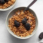 Overhead view of maple granola in a grey bowl and topped with milk and blackberries.