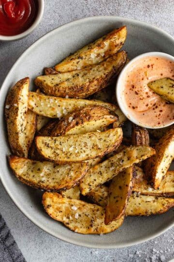 Overhead view of air fryer potato wedges in a grey bowl with dipping sauces off to the sides.