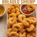 Pinterest image with breaded shrimp piled on a plate with a small bowl of seafood sauce.