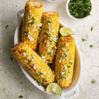 Four ears of air fried corn in a white dish and topped with feta and parsley.