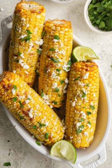 Four ears of air fried corn in a white dish and topped with feta and parsley.