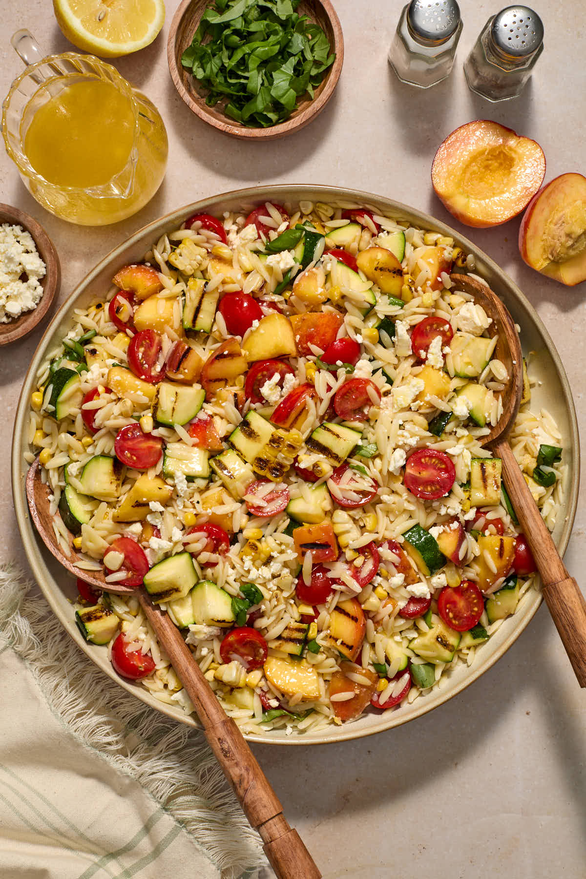 Vegetable orzo salad assembled in a bowl with serving spoons and extra vinaigrette and chopped basil on the side.