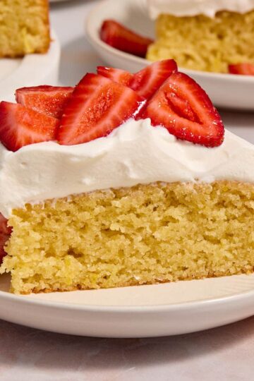 Slice of almond flour lemon cake on a plate and topped with whipped cream and strawberries.