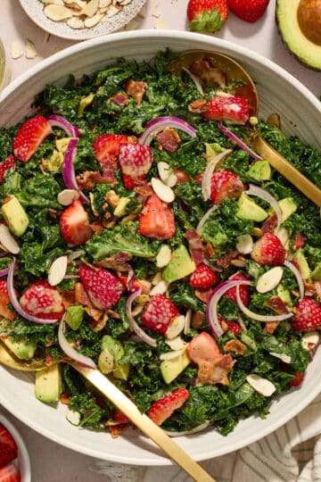 Strawberry kale salad tossed together in a white bowl with extra ingredients surrounding the bowl.