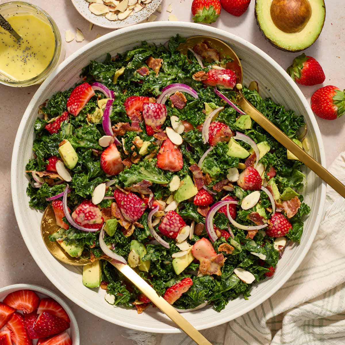 Strawberry kale salad tossed together in a white bowl with extra ingredients surrounding the bowl.