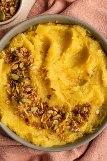 Mashed acorn squash in a bowl on a pink cloth with savoury granola on top.