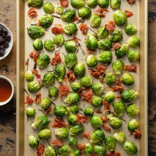 Roasted Brussels sprouts with prosciutto on a baking sheet with dried cranberries and maple syrup off to the side.