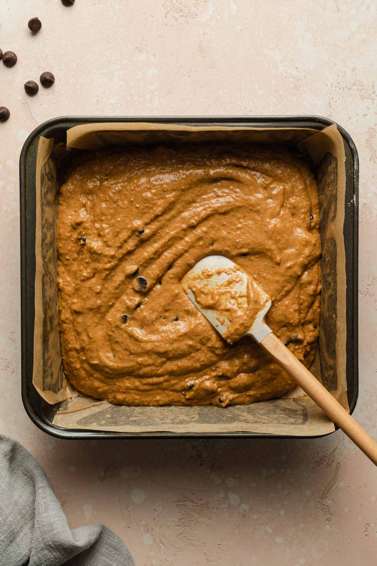 Pumpkin cake batter spread out in a square baking pan with a spatula.