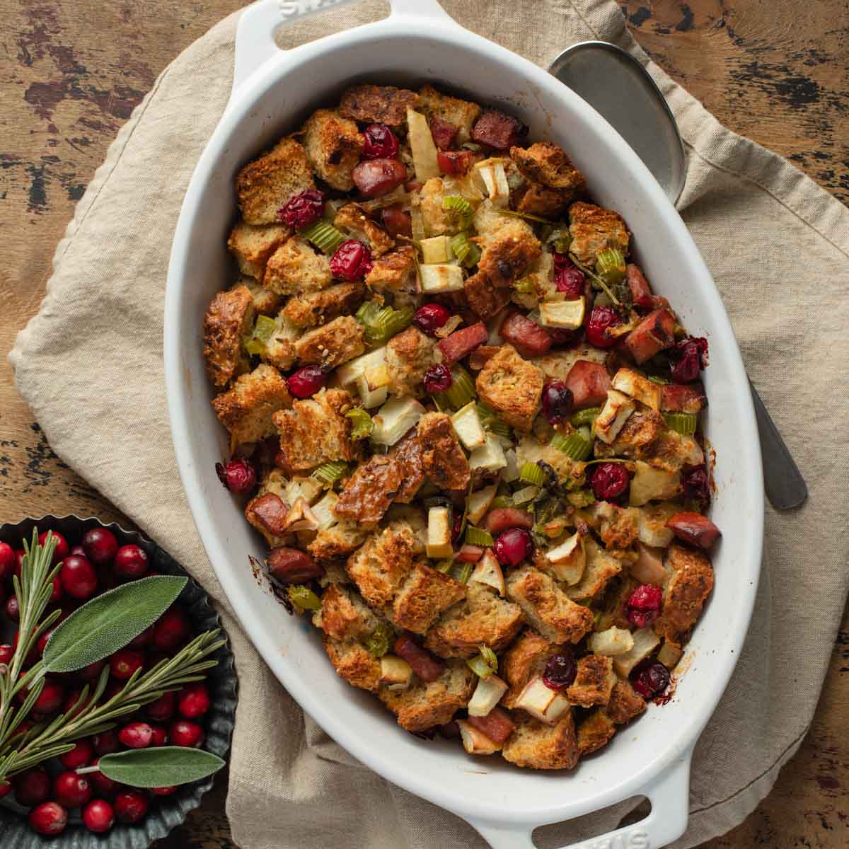 Whole wheat stuffing in a white baking dish with cranberries and sage off to the side.