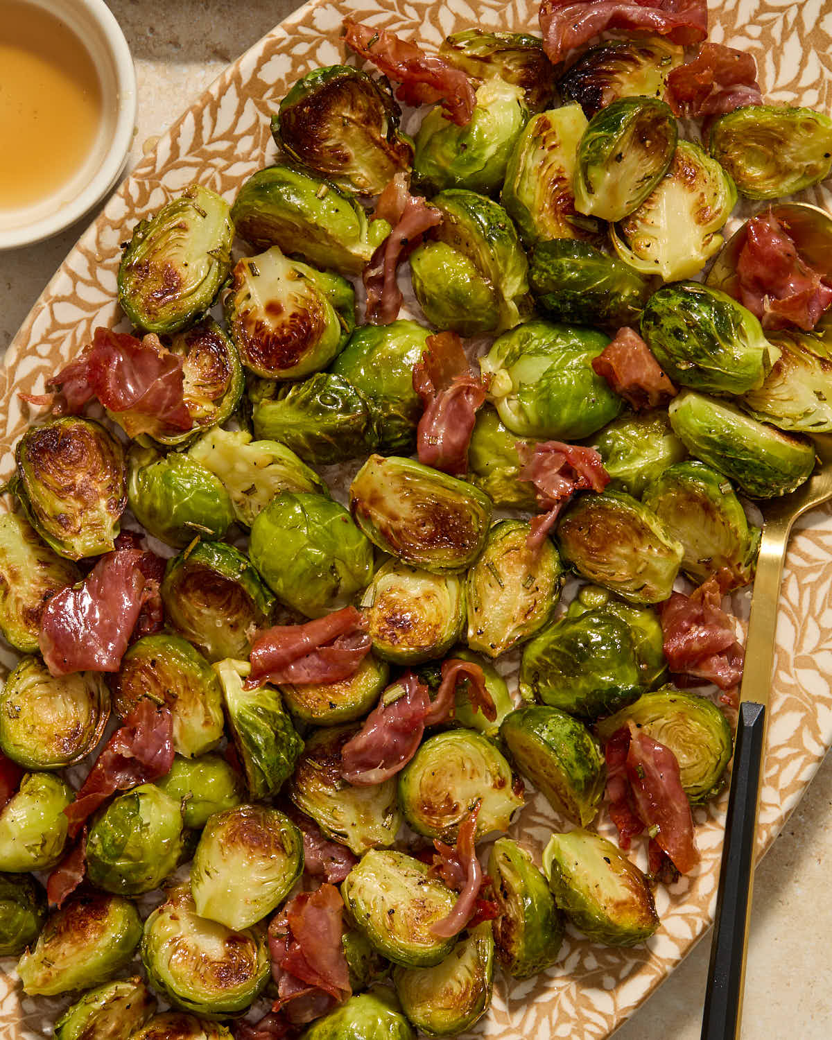Brussels sprouts and prosciutto served on a platter with a serving spoon and maple syrup on the side.