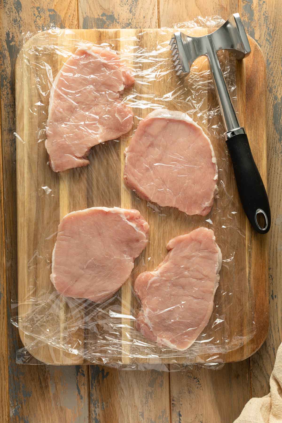 Pork chops on a wooden board in between plastic wrap with a mallet next to them.