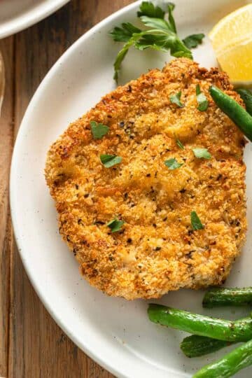 Air fryer pork schnitzel on a white plate with green beans and a lemon wedge.