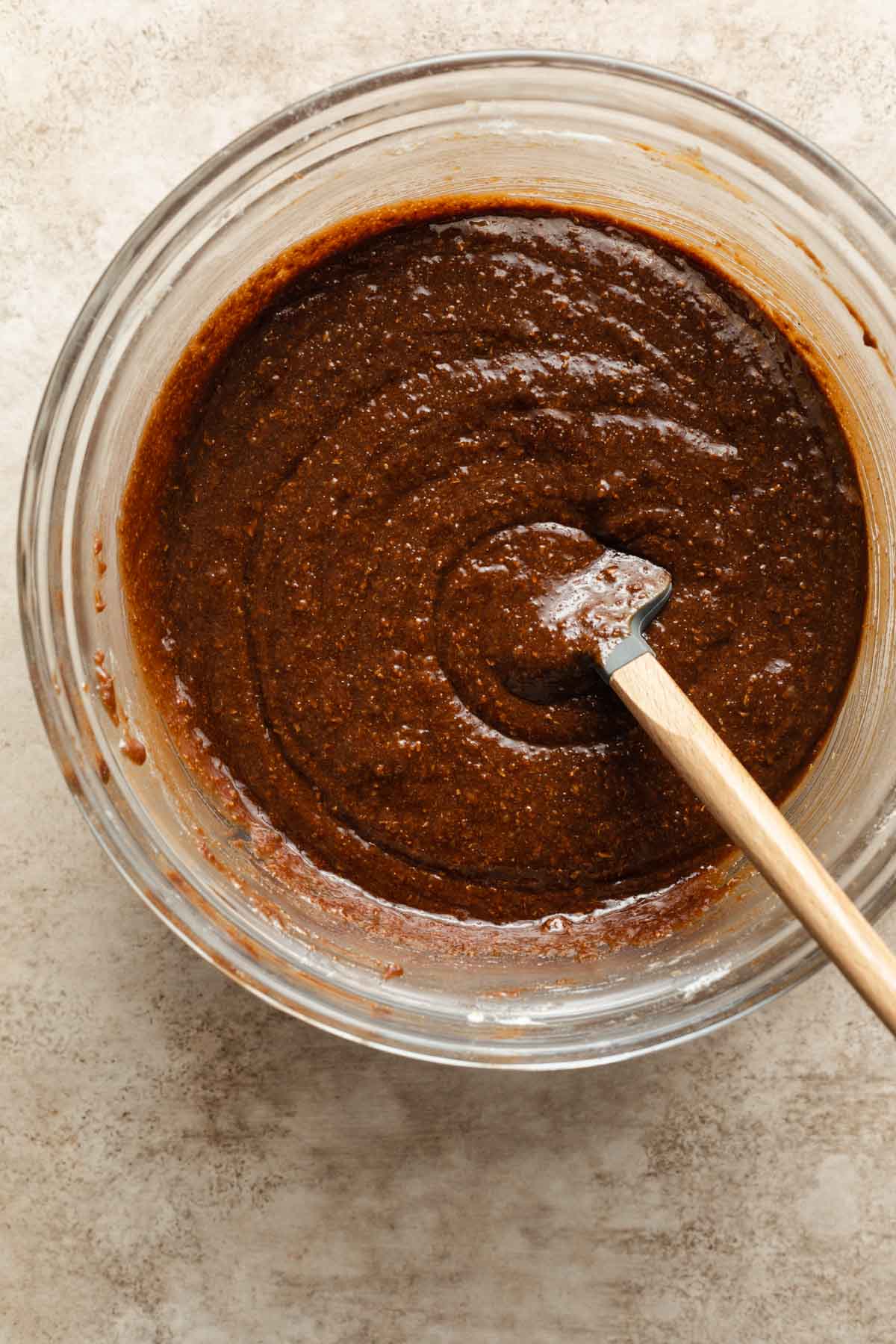 Almond butter brownie batter being stirred in a glass bowl with a spatula.