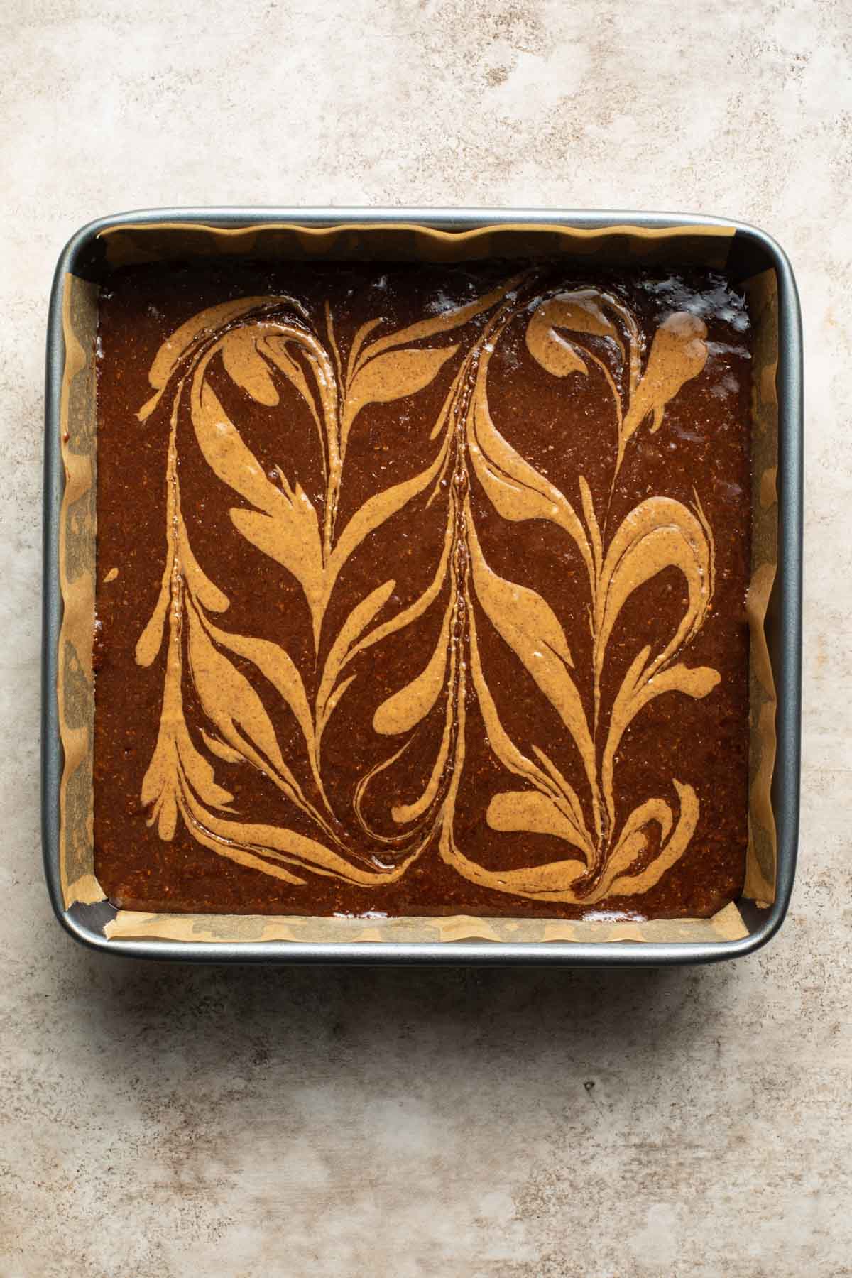 Brownie batter in a square pan with almond butter swirled around on top.