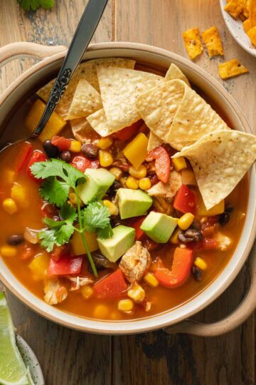 Chicken fajita soup in a beige bowl with a spoon inserted into it and tortilla chips on top.