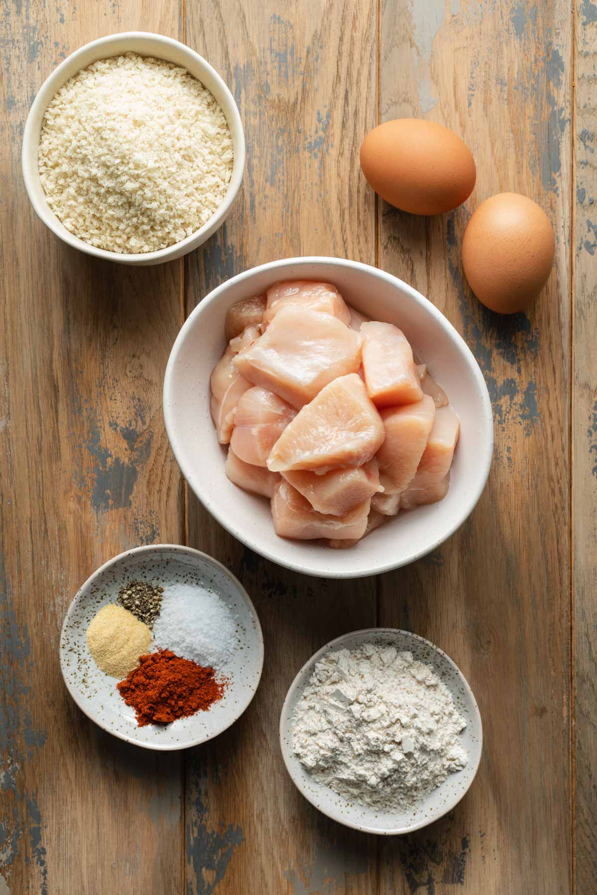 Ingredients to make breaded chicken bites arranged individually on a wooden surface.