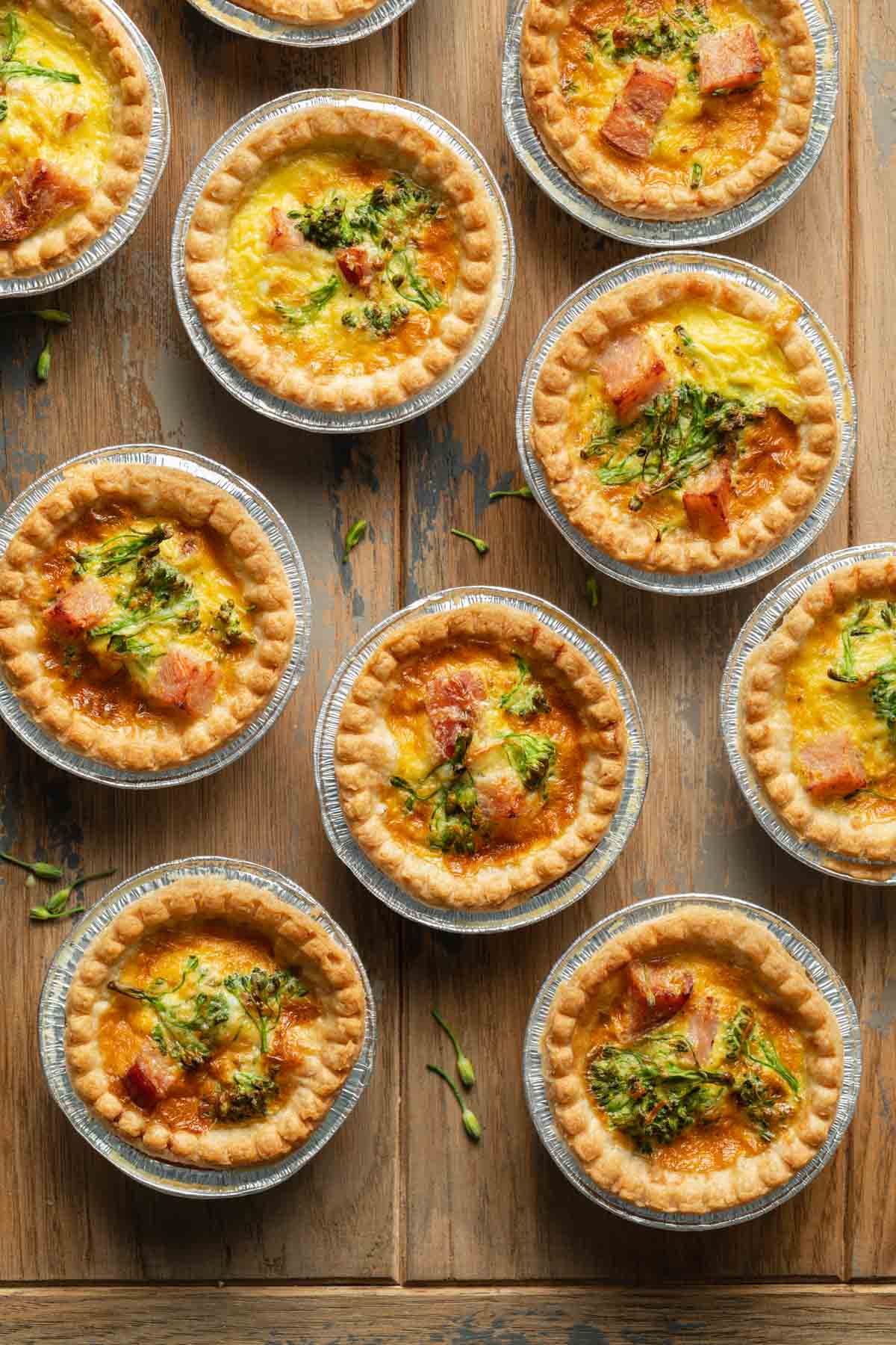 Mini quiches in foil tart shells arranged on a wooden surface.