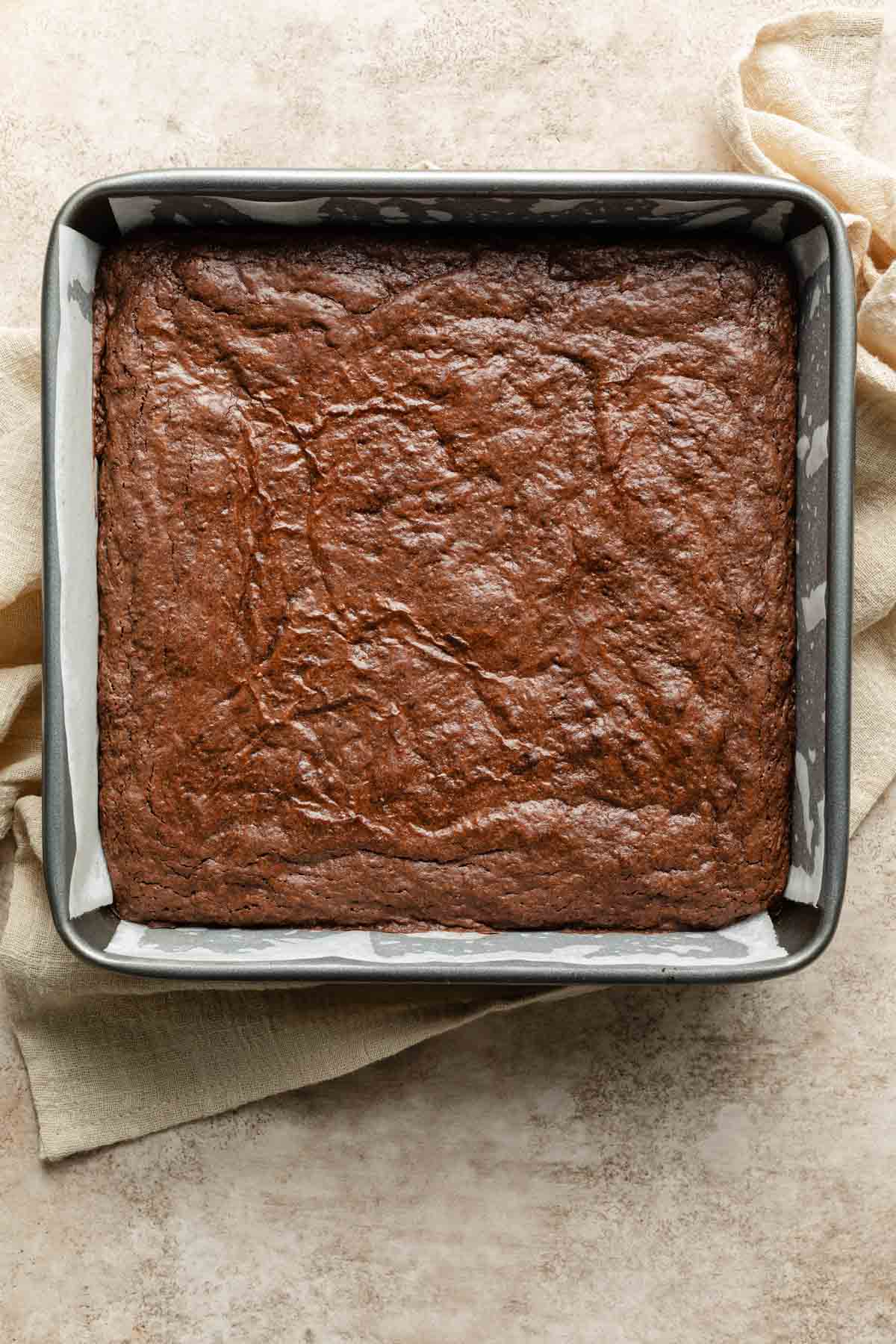 Brownies baked up in a square pan and resting on a beige cloth.