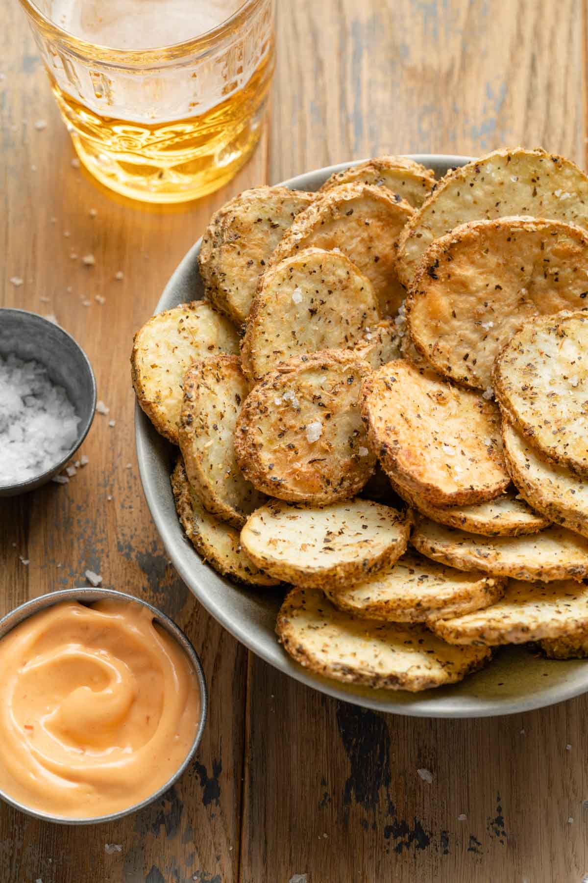 Angled view of potato slices in a grey bowl next to a bowl of dip and a drink in the background.