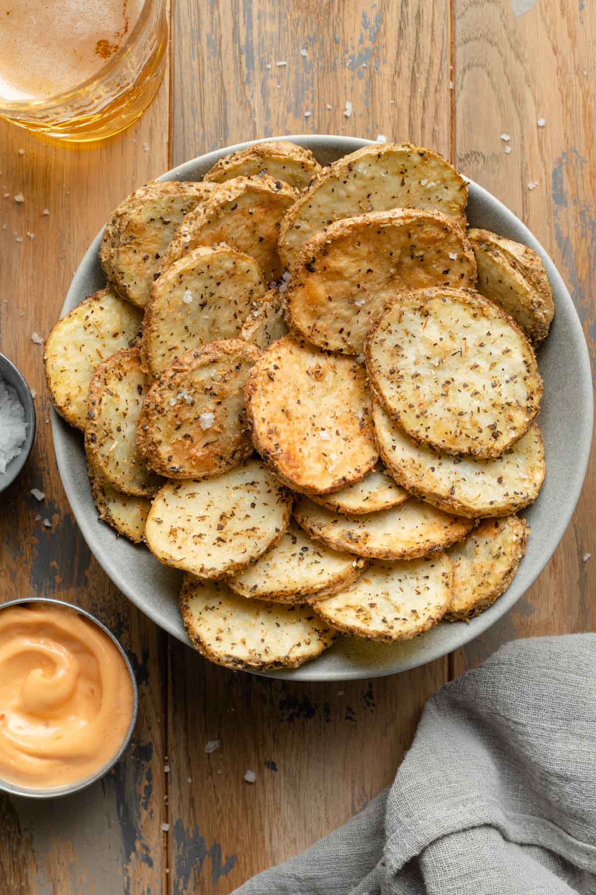 Overhead view of air fryer potato slices in a bowl on wooden backdrop next to dip, a drink and a grey napkin.