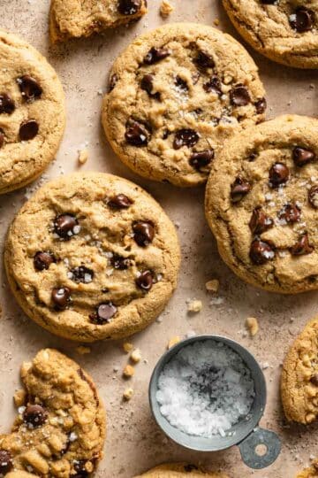Oat flour chocolate chip cookies arranged on a beige surface next to a measuring cup of flaky sea salt.