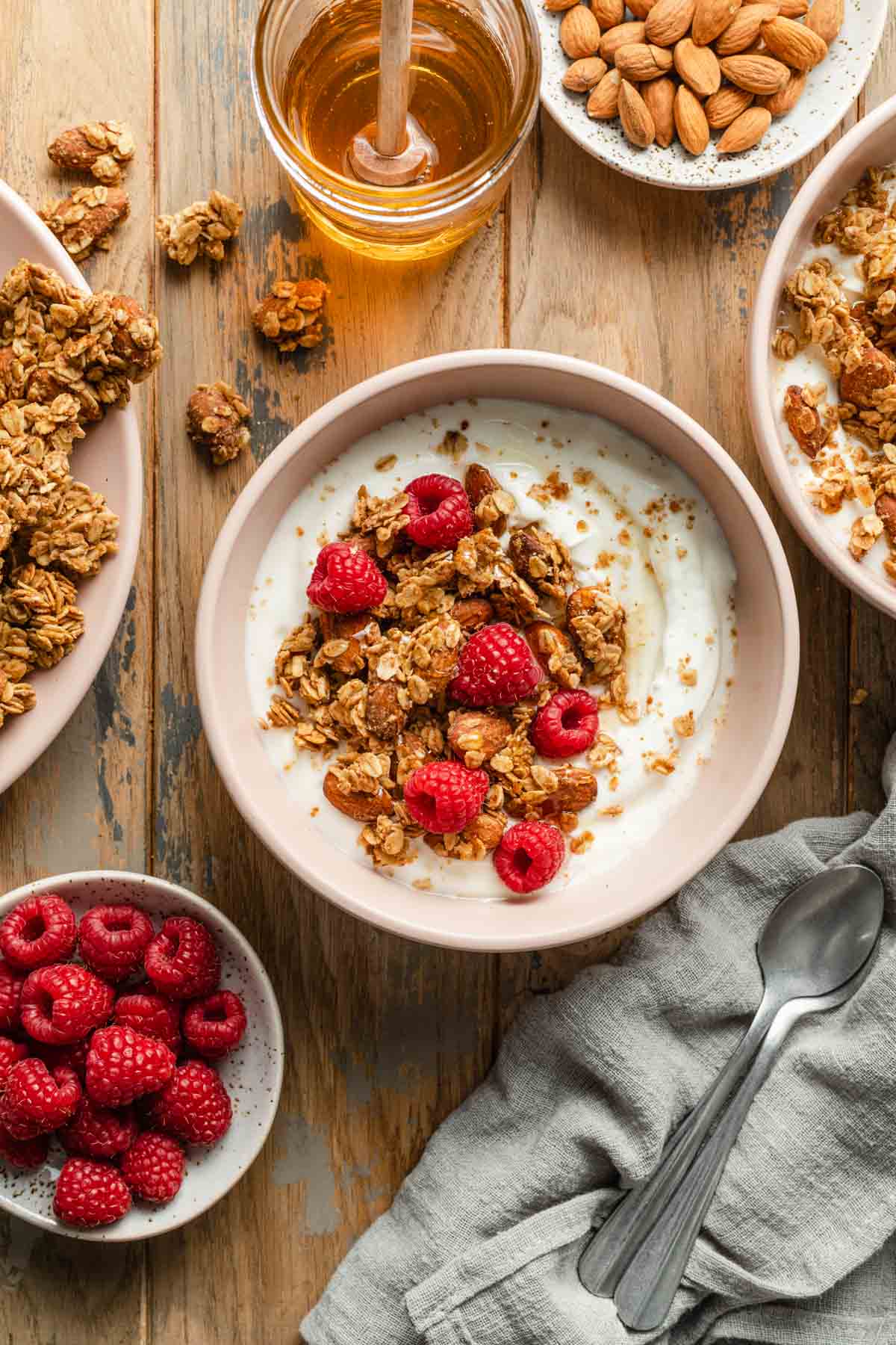 A bowl of yogurt, raspberries and granola on a wooden table surface with raspberries, honey and granola off to the sides.
