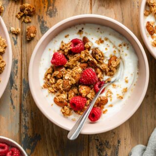 Overhead view of honey almond granola with yogurt and raspberries in a pink bowl with a spoon.