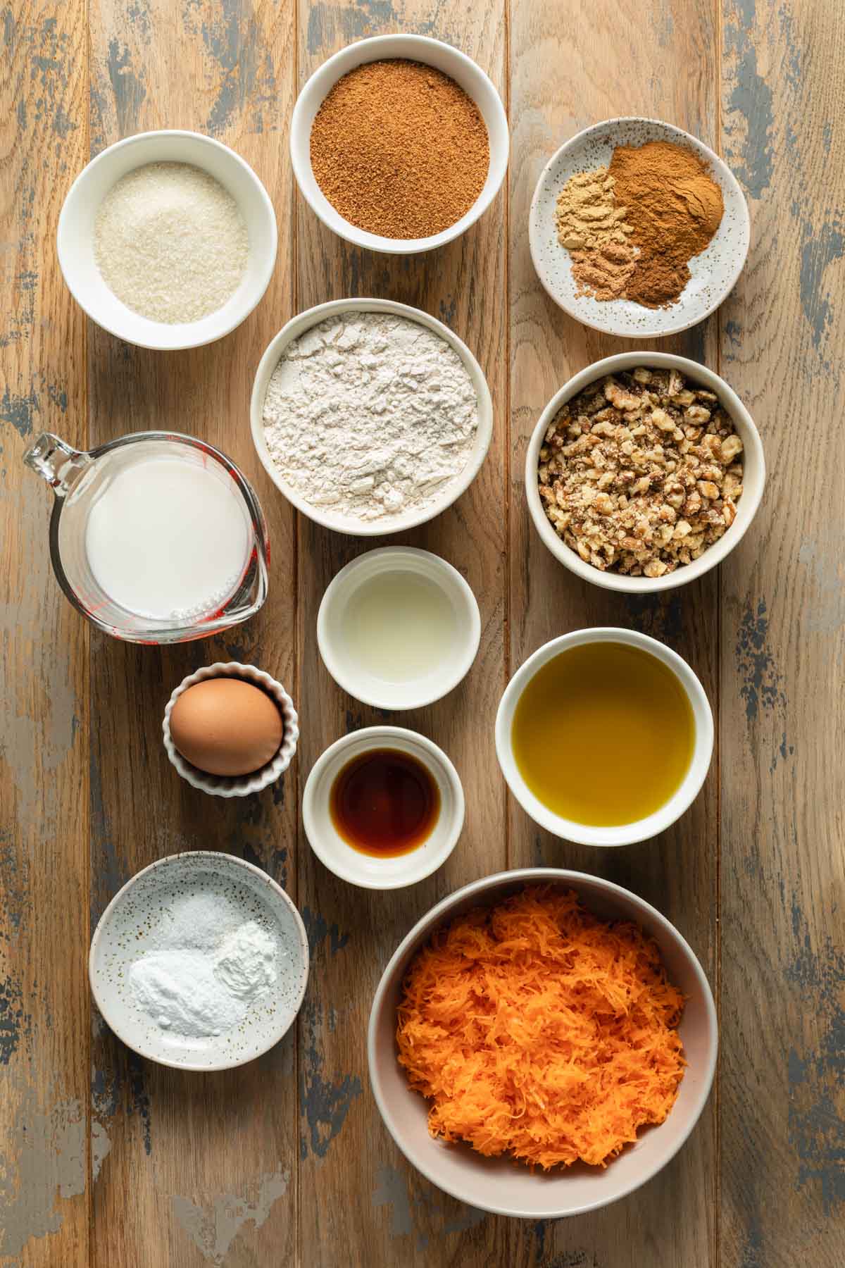Ingredients to make this small carrot cake recipe arranged individually on a wooden table surface.
