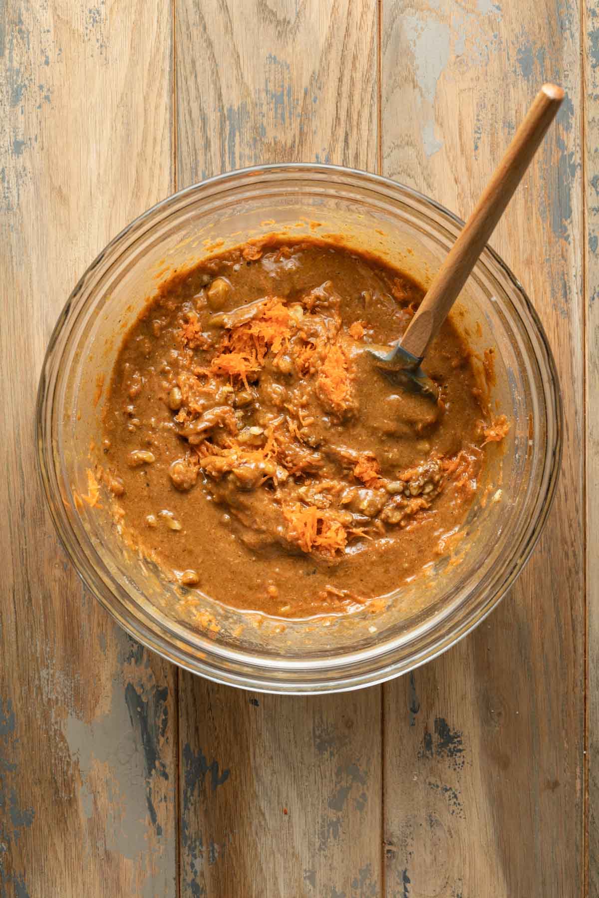 Carrot cake batter in a glass bowl with carrots and walnuts mixed in with a spatula.