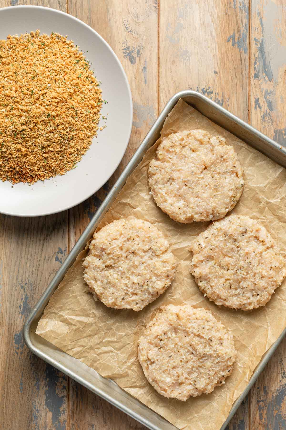 Chicken patties arranged on parchment paper next to a bowl of seasoned breadcrumbs.