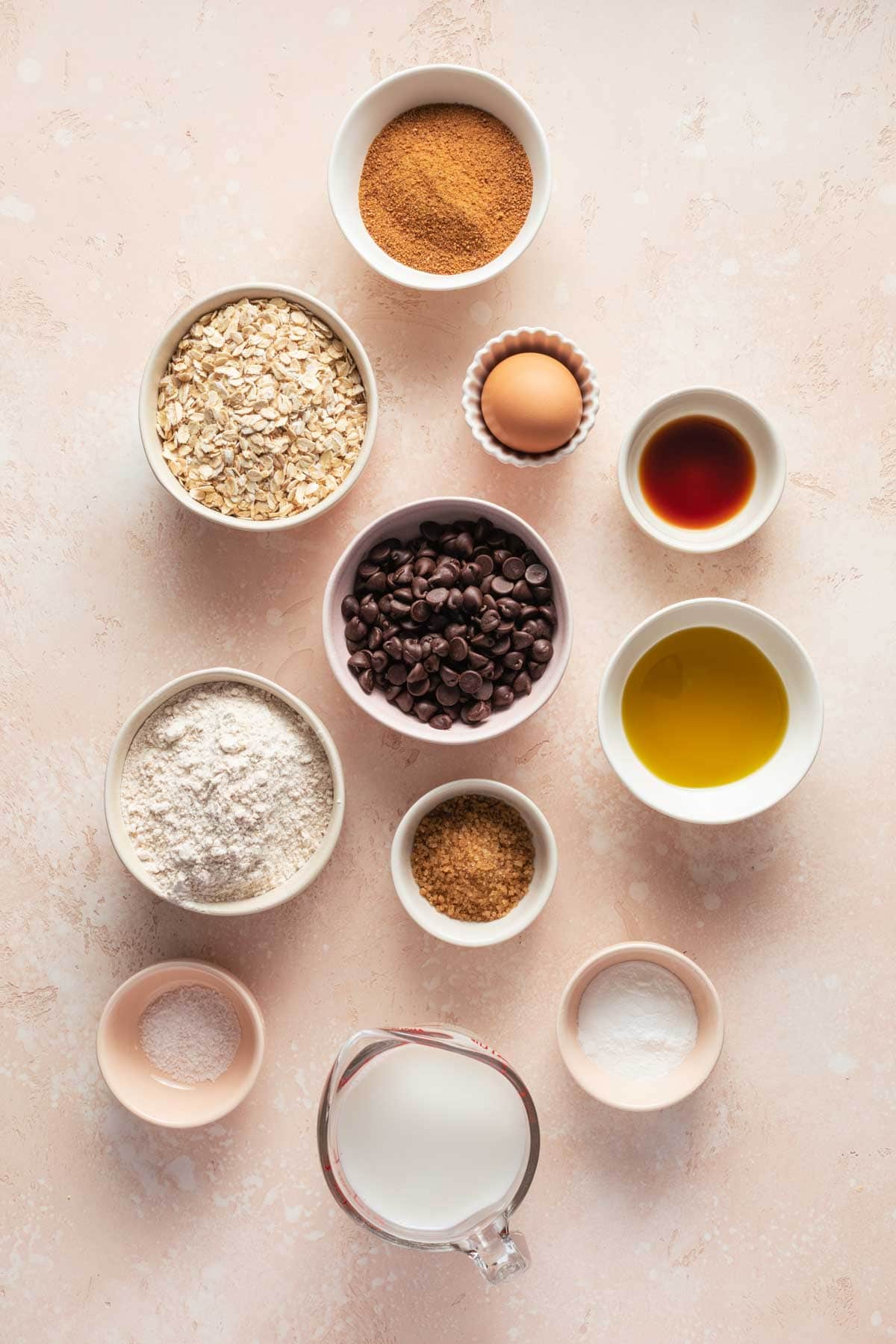 Ingredients to make air fryer muffins arranged in individual dishes on a pink surface.