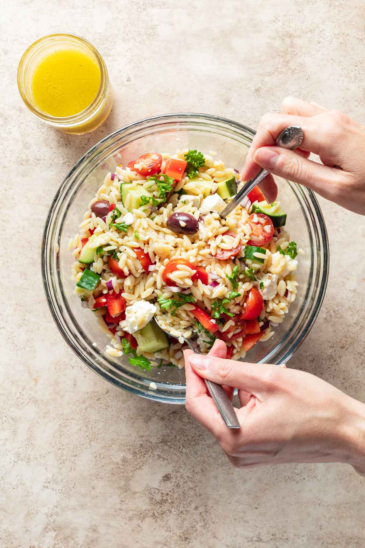 Orzo salad being tossed together in a glass bowl with a jar of lemon vinaigrette on the side.