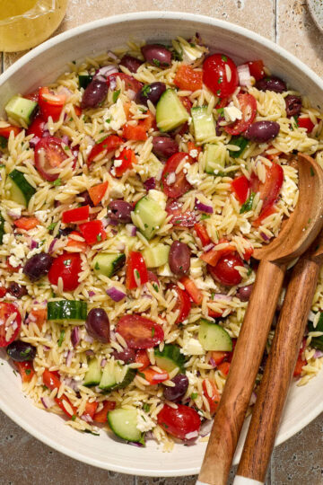 Greek orzo pasta salad in a white bowl with wooden salad servers and feta, lemon dressing and parsley on the side.