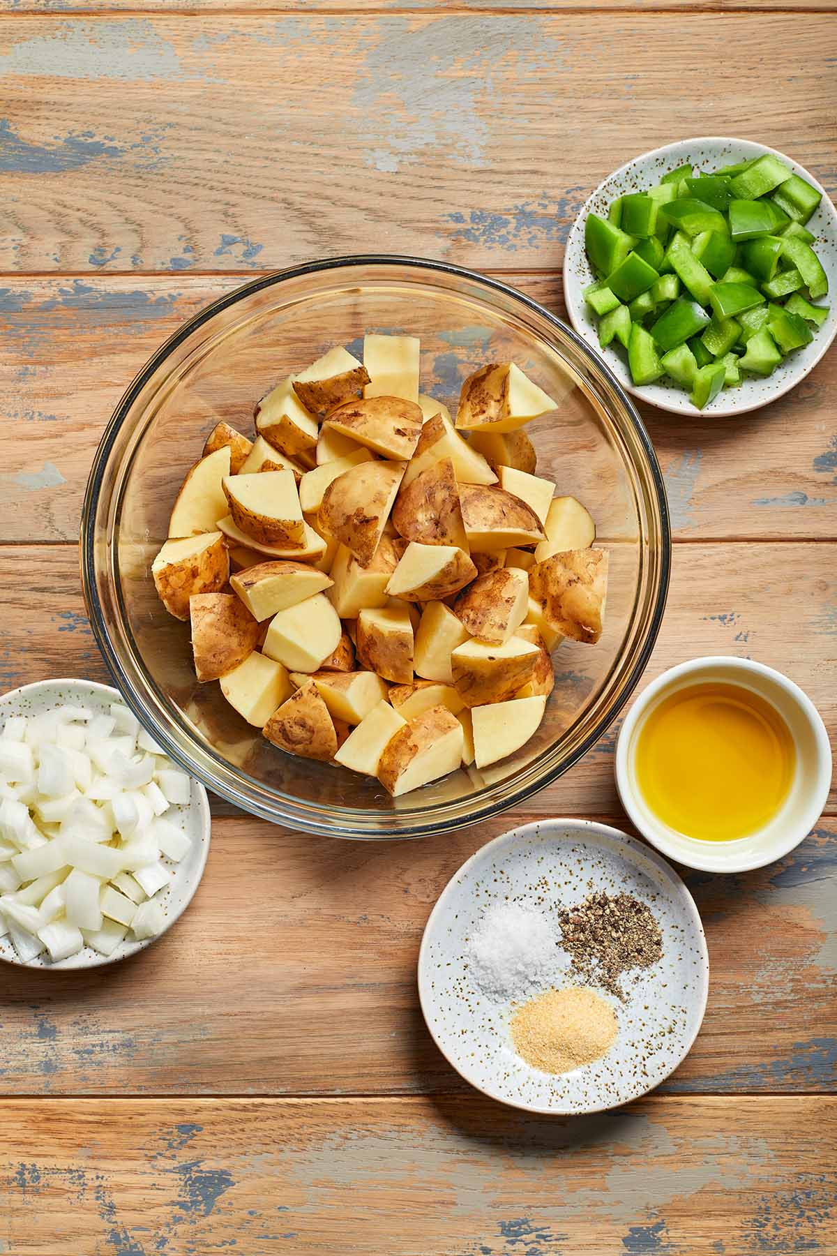 Ingredients to make air fryer home fries arranged in individual dishes on a wooden surface.