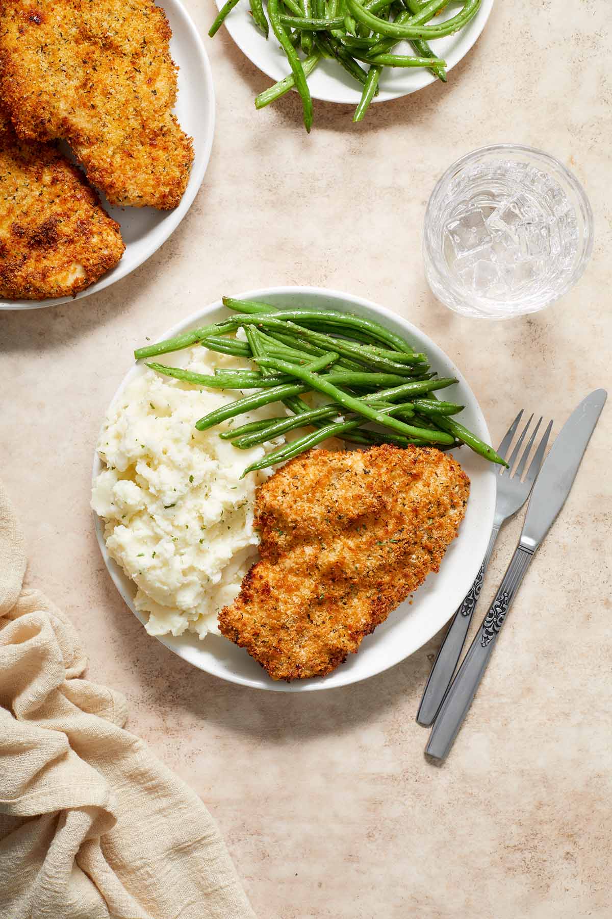 Overhead view of chicken schnitzel, mashed potatoes and green beans on a plate with plates of chicken and green beans off to the side.