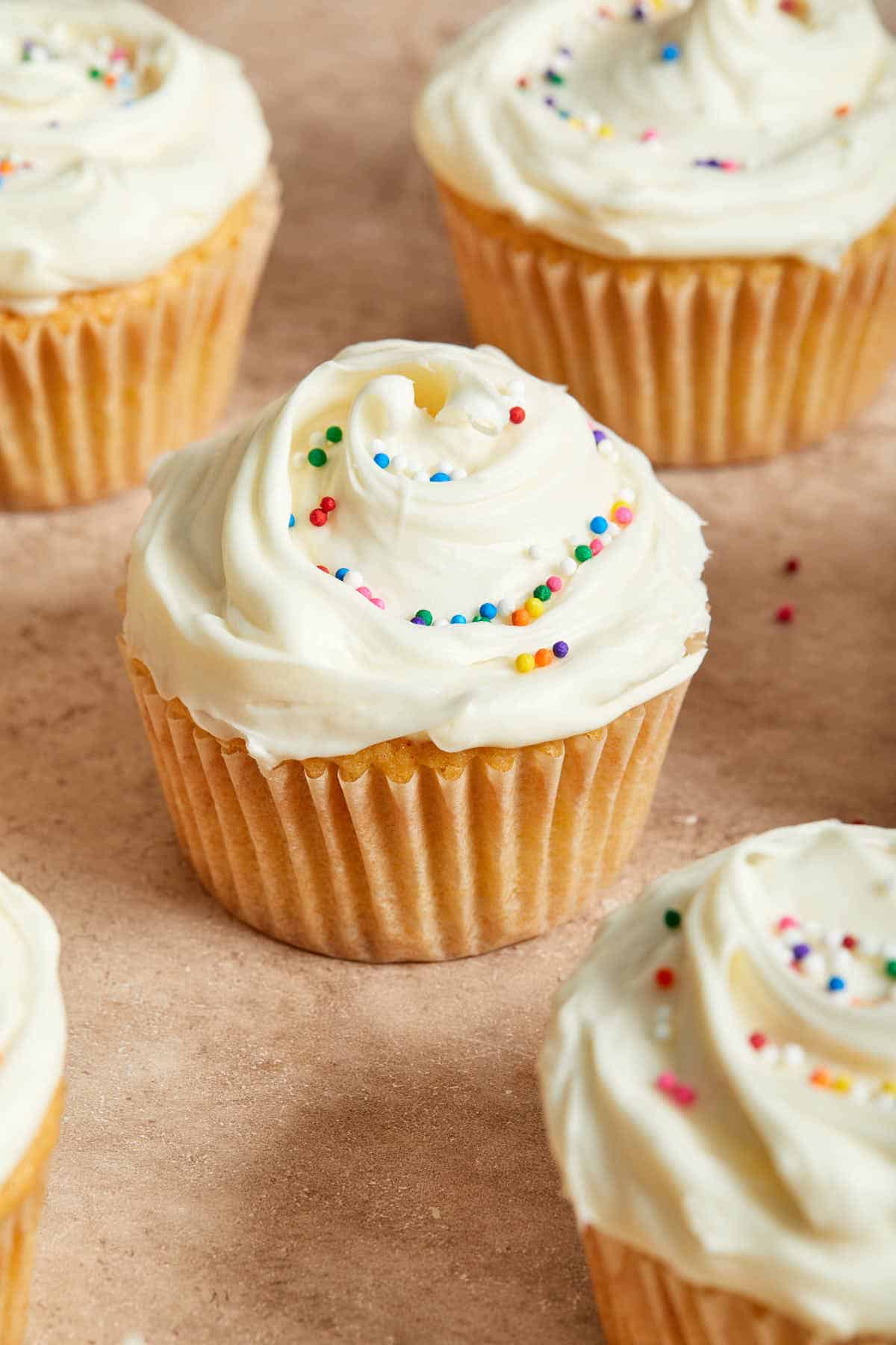 Frosted cupcakes with sprinkles, arranged on a beige coloured surface.