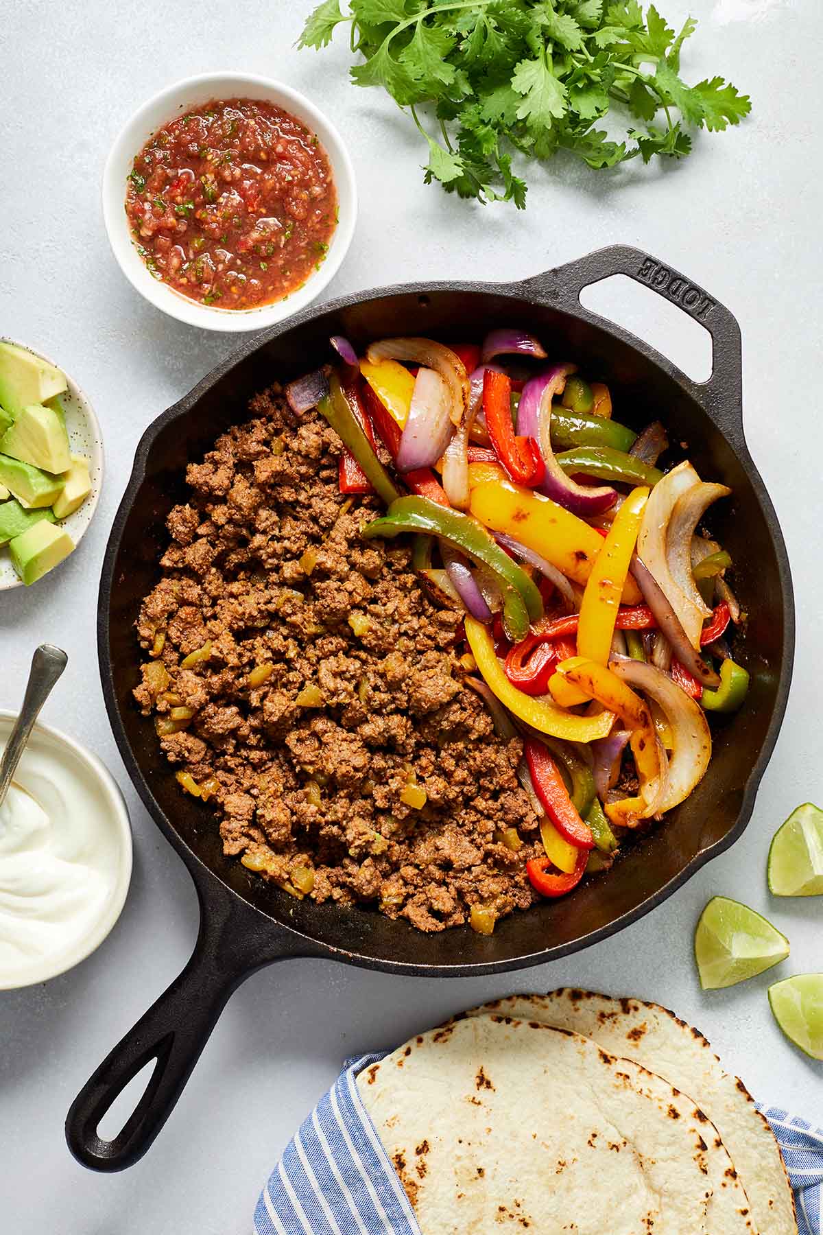 Ground beef, peppers and onions in a cast iron pan with fajita toppings surrounding it.