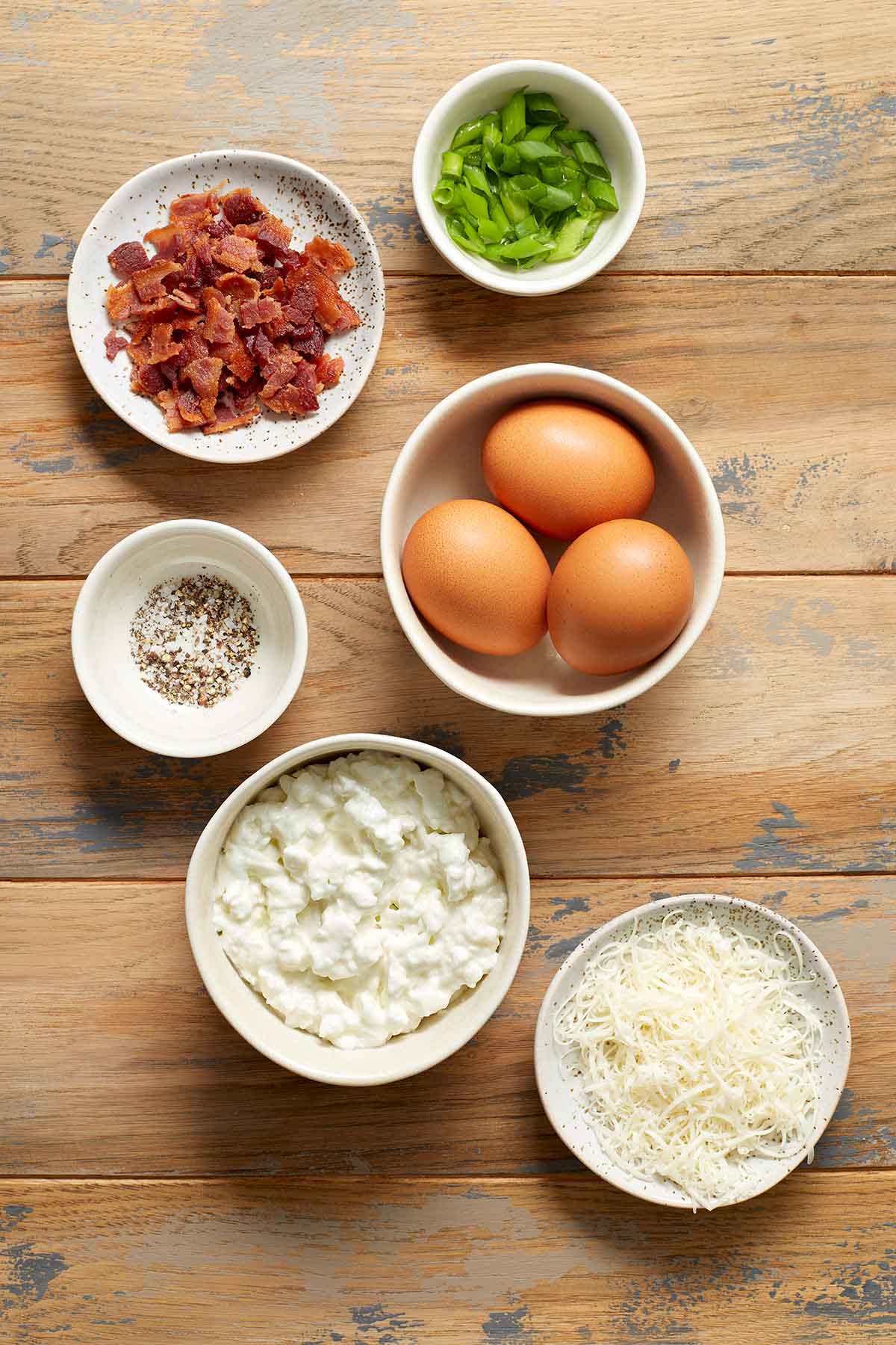 Ingredients to make air fryer egg bites arranged in individual bowls on a wooden surface.