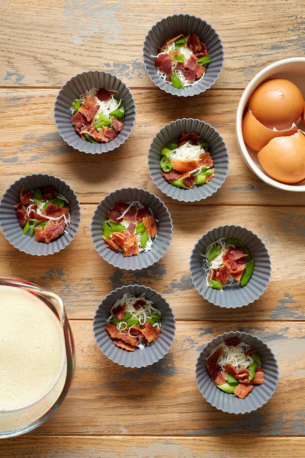 Silicone muffin cups filled with bacon, cheese and green onion with the egg mixture in a cup on the side.